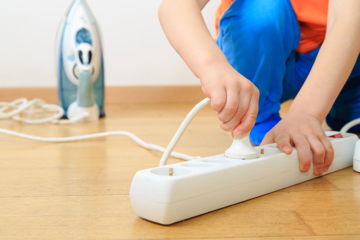 A child is putting the plug of flat iron into a power outlet on a wooden floor. 