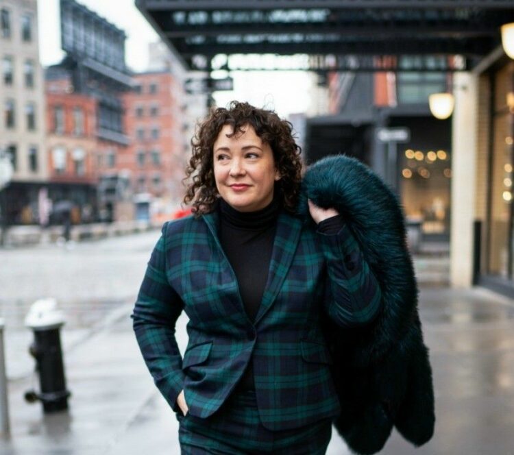 A woman with curly hair wearing a plaid blazer holds a green fur coat over her shoulder on a city street.