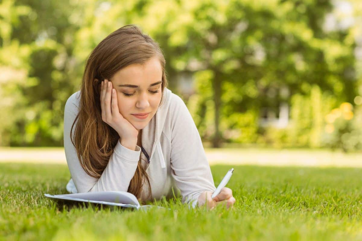Woman lying on the grass and jotting down journal ideas for teens in a notebook.