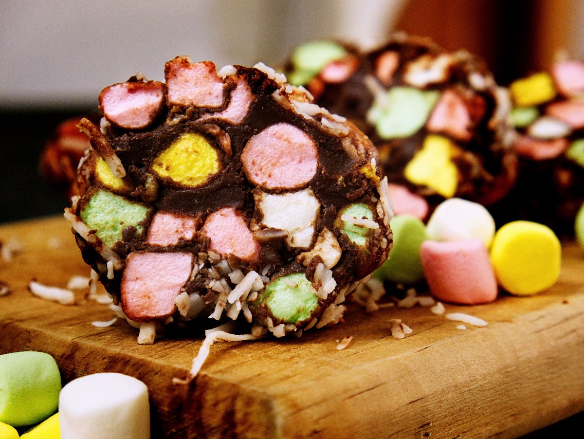 Colorful marshmallow treat coated with chocolate and sprinkles on a wooden cutting board.