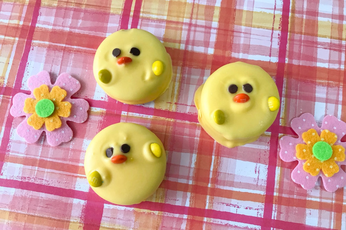 Four decorated cookies resembling chicks, with two floral cookies, on a checkered pink background.