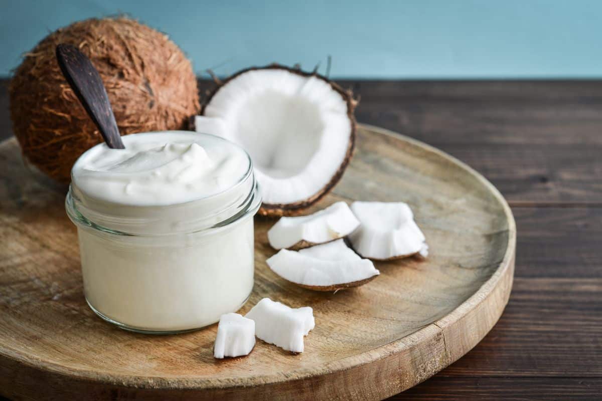 Jar of coconut cream with a spoon on a wooden plate next to fresh coconut pieces.
