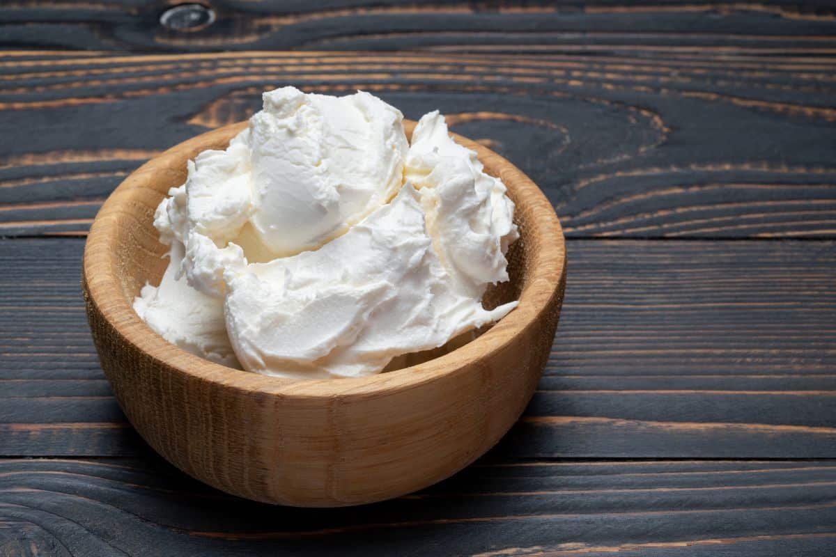 A wooden bowl filled with mascarpone, a replacement for ricotta cheese, on a dark wooden background.