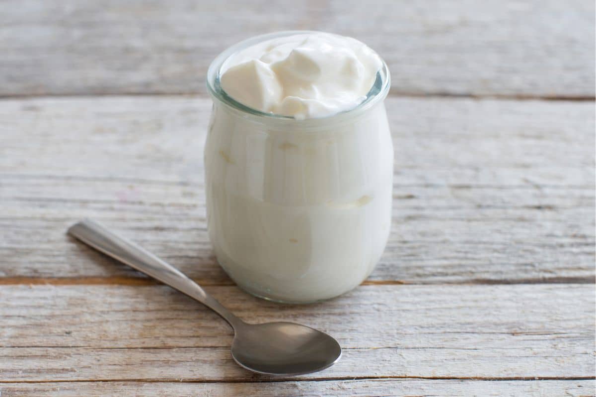 A jar of greek yogurt, a replacement for Ricotta cheese, with a spoon on a wooden surface.