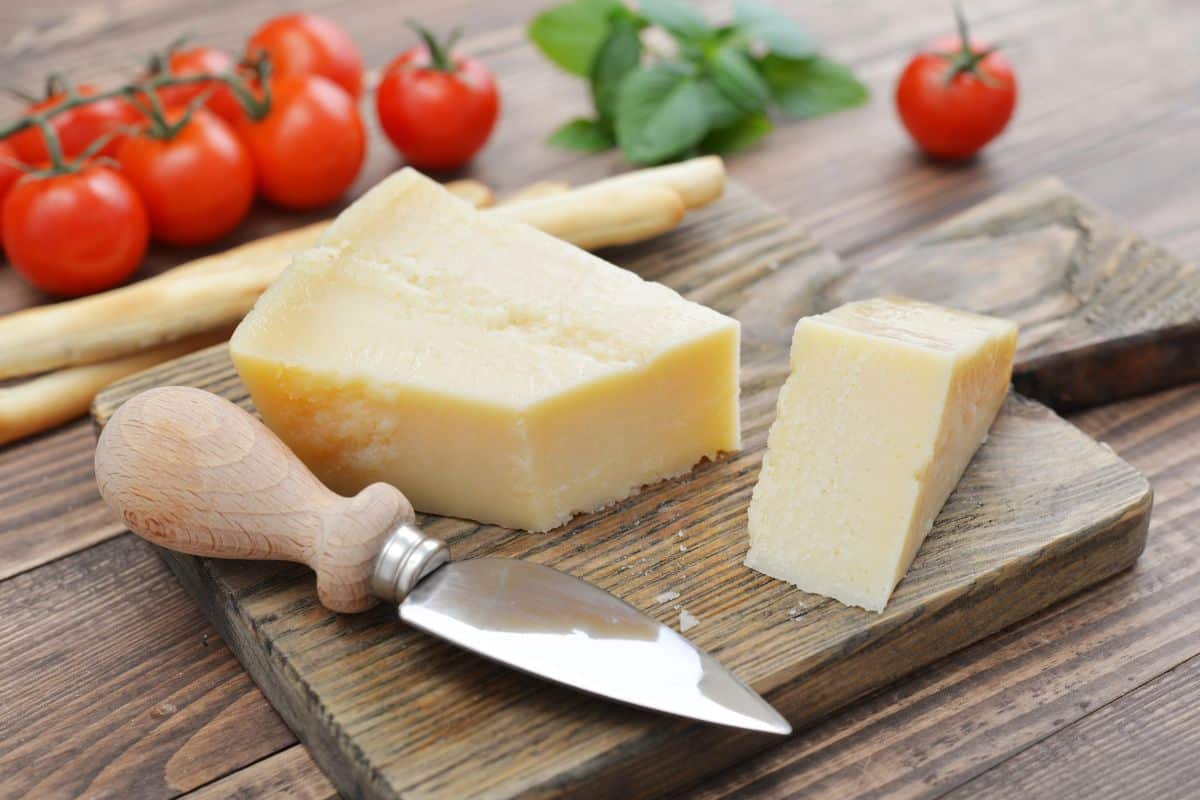 Wedge of parmesan cheese with a cheese knife on a wooden board, accompanied by tomatoes and basil.