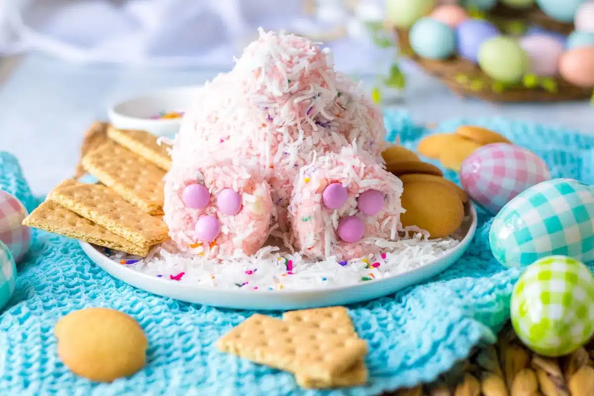 A festive easter dessert shaped like an egg and coated with pink shredded coconut, adorned with candy eggs and served with cookies and graham crackers on a cheerful, springtime table setting.