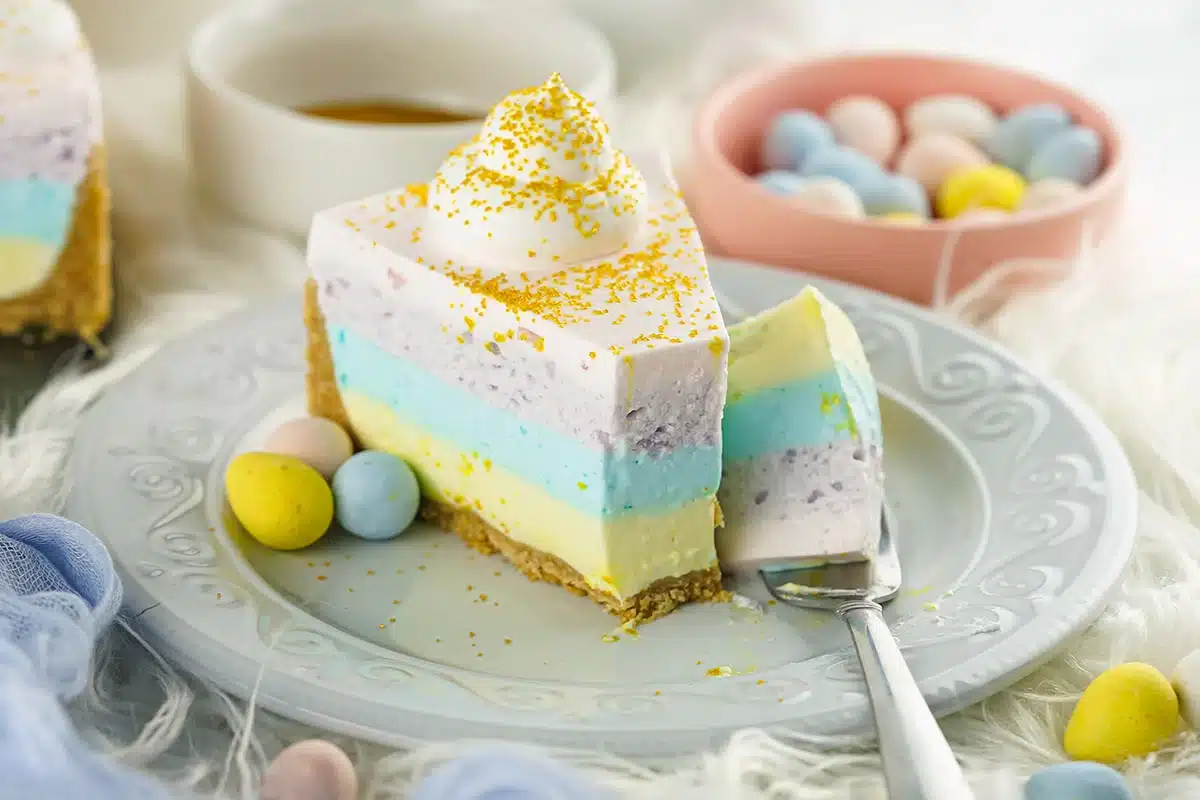 A colorful layered easter cheesecake with a lemon garnish on a plate, accompanied by pastel candy eggs and a cup of tea.