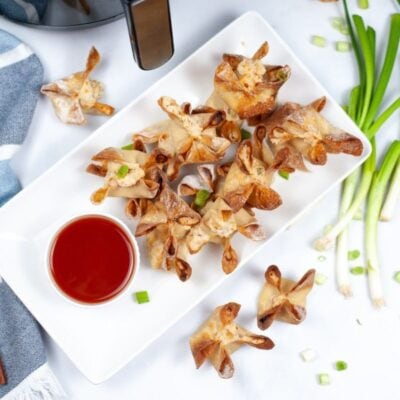 Plate of crispy wontons with a bowl of red dipping sauce, surrounded by green onions and chopsticks on a marble surface.