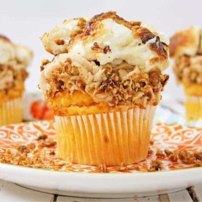 A cupcake topped with crispy shredded chicken and melted cheese sits elegantly on a decorative plate, reminiscent of unique Thanksgiving desserts. Two similar cupcakes are in the background.