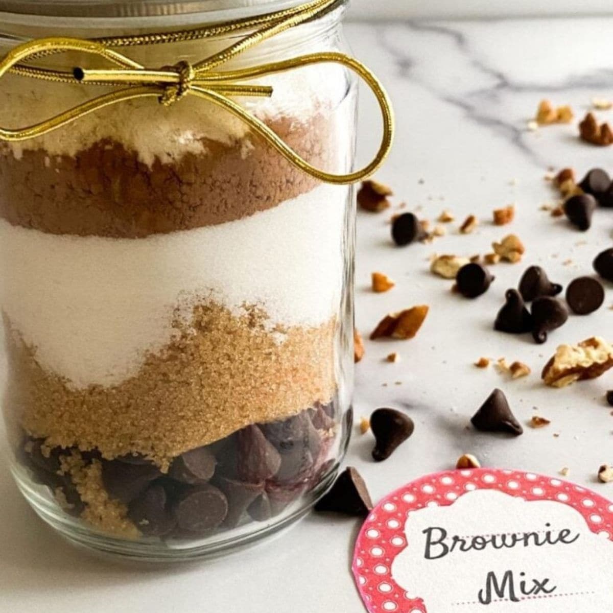 A glass jar filled with layered brownie mix ingredients, topped with a gold ribbon and a “Brownie Mix” tag.