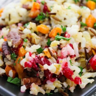 Close-up of a colorful rice dish with wild rice, cranberries, nuts, carrots, and fresh parsley on a dark plate.