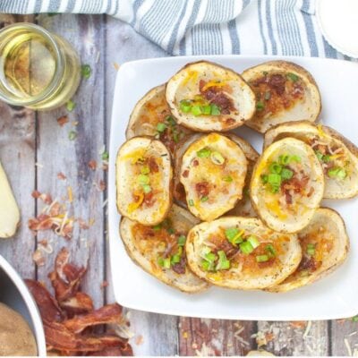 A plate of potato skins topped with cheese, bacon bits, and chopped green onions on a wooden table with drinks and a striped napkin.