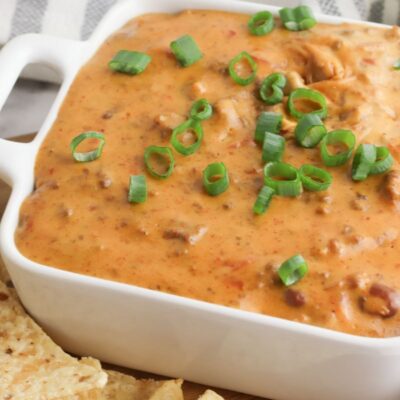 A square white bowl filled with creamy queso dip, topped with sliced green onions, surrounded by tortilla chips on a table.