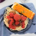 A plate of spaghetti topped with vegetarian eggplant meatballs and savory tomato sauce, accompanied by a slice of cheesy garlic bread, rests on a blue cloth.