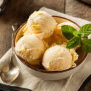 A bowl of vanilla ice cream, made even creamier with sweetened condensed milk, is topped with a mint garnish and comes with an accompanying spoon on a wooden surface.