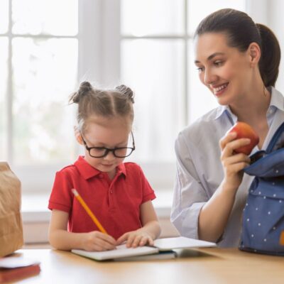 A woman holding an apple smiles while a child in glasses writes in a notebook. A blue backpack and a brown paper bag are on the table.