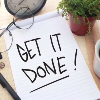 Notebook with "GET IT DONE!" written on it, resting on a table with a clock, glasses, pen, potted plant, and cup of coffee.