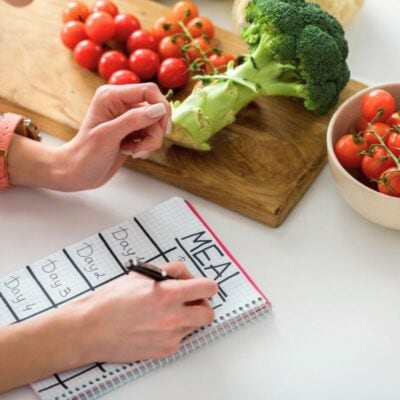 Person writing a meal plan in a notebook at a table with tomatoes, broccoli, and cauliflower.
