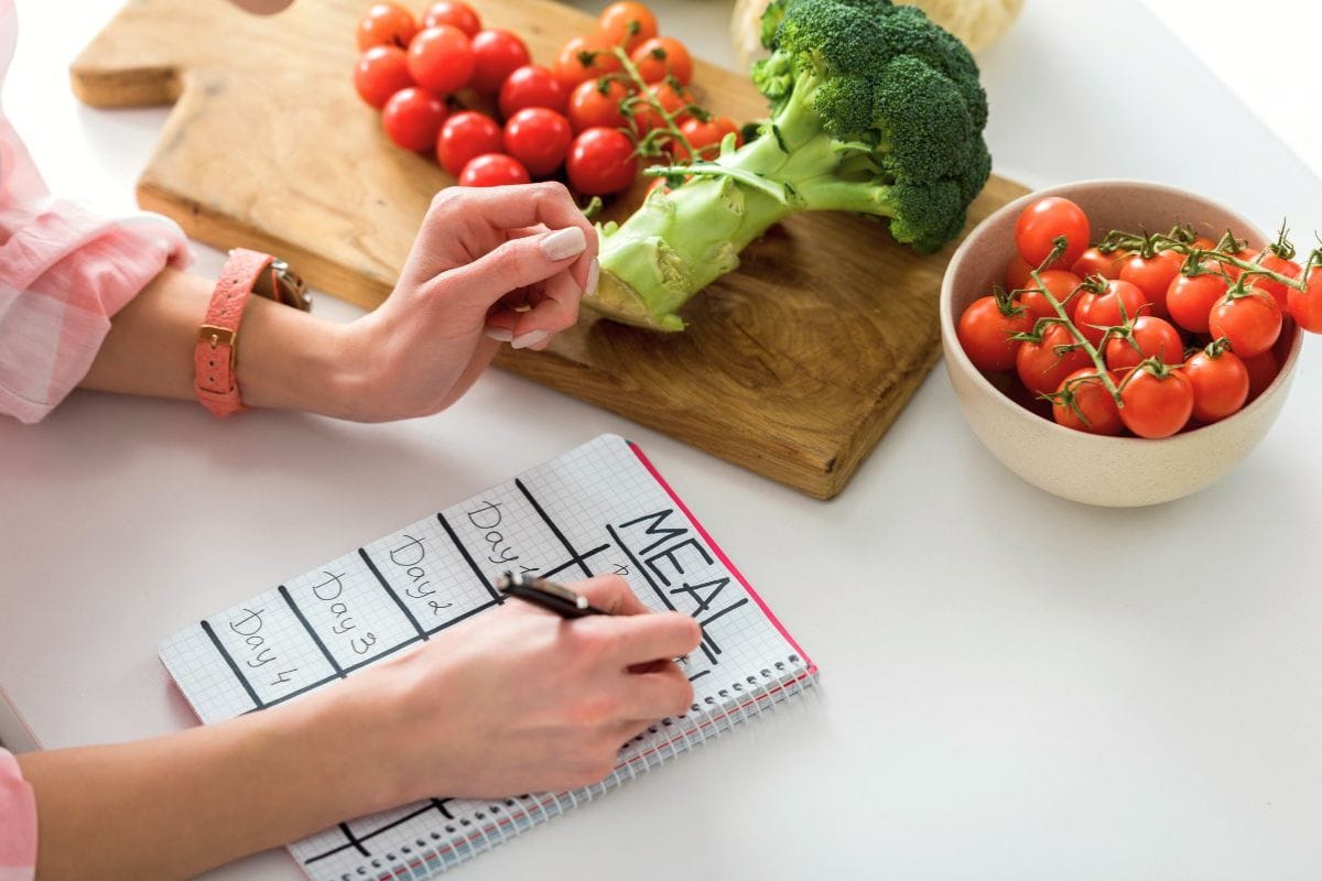 Person writing a meal plan in a notebook at a table with tomatoes, broccoli, and cauliflower.