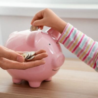 A child places a coin into a pink piggy bank, while an adult holds more coins in their hand.