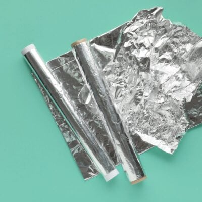 Two rolls of aluminum foil with an unrolled sheet, on a teal background.