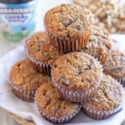 A basket lined with a white cloth holds a stack of muffins with visible chocolate chunks. An ice cream container and chopped nuts are in the background.