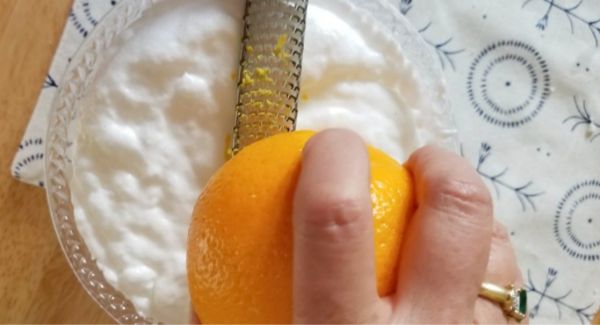 A hand is grating an orange over a bowl of whipped egg whites with a fine grater on a patterned cloth.