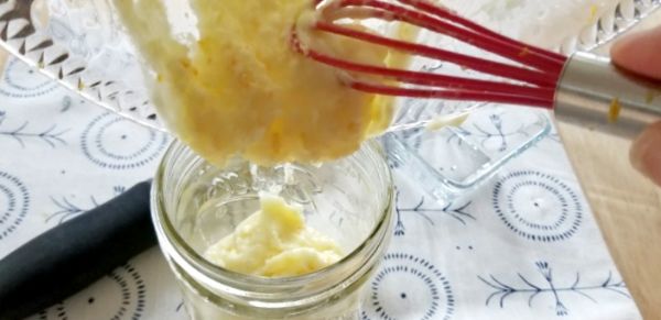 A hand uses a red whisk to transfer a creamy mixture from a glass bowl into a small glass jar on a patterned cloth.