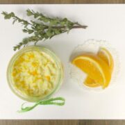 A small jar of creamy dessert with orange zest, a sprig of greenery, and three orange slices on a doily, arranged on a white surface.