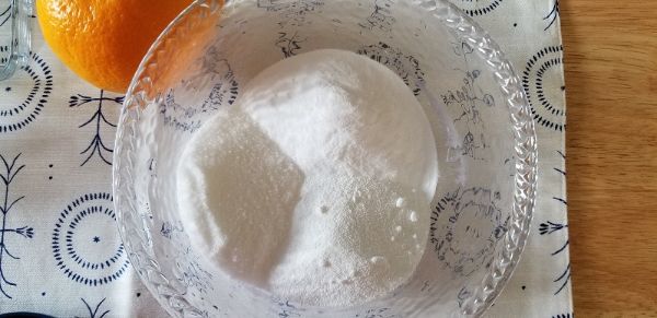 A clear glass bowl filled with white granulated sugar sits on a patterned tablecloth next to an orange.