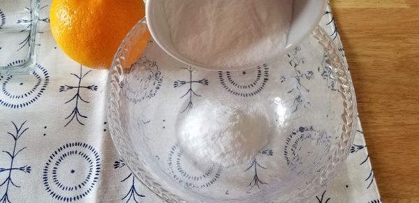 A clear glass bowl with white powder being poured in, next to a whole orange, on a patterned tablecloth.