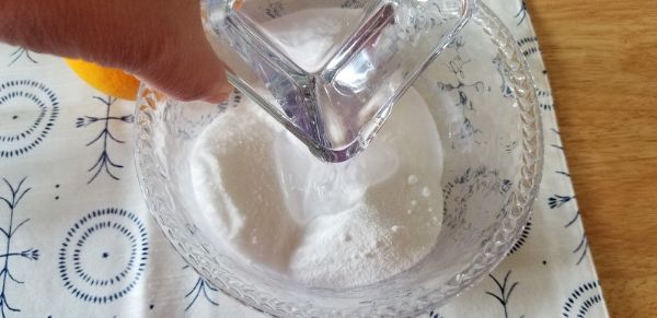 A hand pours water from a glass pitcher into a bowl containing white powder on a patterned cloth.