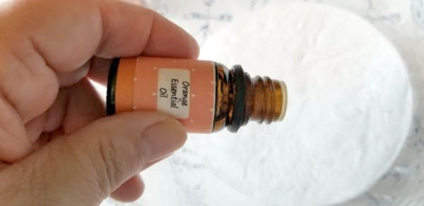 A hand holds a small bottle labeled "Orange Essential Oil" above a white surface.
