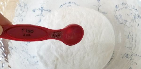 A red 1 teaspoon measuring spoon is held above a bowl filled with white powder, likely flour or sugar.