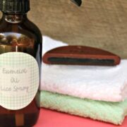 A brown bottle labeled "Essential Oil Lice Spray" sits beside a nit comb and two folded towels on a pink surface.