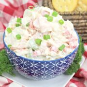A blue patterned bowl filled with creamy salad topped with chopped green onions, placed on a white plate with parsley, on a red and white checkered cloth. Crackers are in the background.