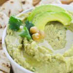 A bowl of avocado hummus topped with chickpeas, cilantro, and a drizzle of olive oil, served with pita bread.