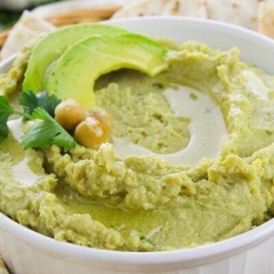 A bowl of creamy avocado hummus garnished with avocado slices, chickpeas, cilantro, and a drizzle of oil, surrounded by pieces of flatbread.