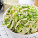 A bowl of noodle salad with shredded vegetables, chopped green onions, and a hint of california roll cucumber salad vibes, placed on a checkered cloth.