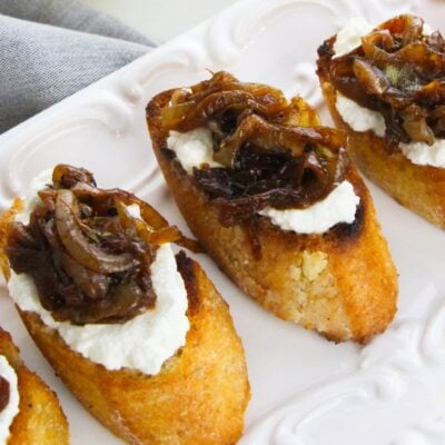 Toasted baguette slices topped with creamy ricotta and sweet caramelized onions, these caramelized onion and goat cheese bites are artfully arranged on a white plate.