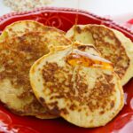 A close-up of three golden brown cottage cheese oat pancakes on a red plate with syrup being poured over them.