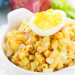A bowl of deviled egg pasta salad with chopped celery and red onion, topped with a halved boiled egg showing its vibrant yellow yolk.