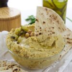 A glass bowl of dill pickle hummus topped with chickpeas and chopped pickles, served with a triangular piece of pita bread and extra pita on the side.