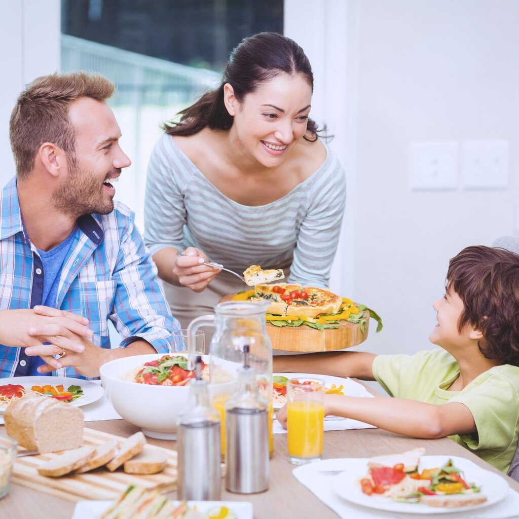 A woman serves pizza to a smiling boy at a dining table with a man and various dishes, including salad, juice, and bread—perfect for a relaxed summer meal plan.