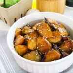 A white bowl filled with gochujang Brussels sprouts, roasted to perfection and topped with sesame seeds, sits on a striped cloth.