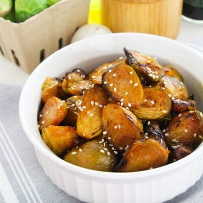 A white bowl filled with gochujang Brussels sprouts, roasted to perfection and topped with sesame seeds, sits on a striped cloth.