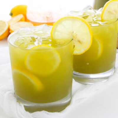 Two glasses of iced matcha lemonade garnished with lemon slices sit on a white surface, with cut lemons and a beverage container in the background.