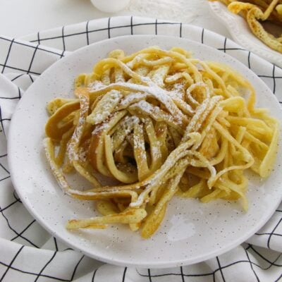 A plate of funnel cakes dusted with powdered sugar on a white speckled dish, set on a white cloth with a black grid pattern—perfect for those who love creative spaghetti pancakes recipes.