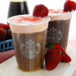 Two Starbucks cold drinks topped with strawberry cold foam and garnished with strawberries, surrounded by fresh berries and a drink in the background.
