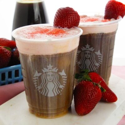 Two Starbucks cold drinks topped with strawberry cold foam and garnished with strawberries, surrounded by fresh berries and a drink in the background.